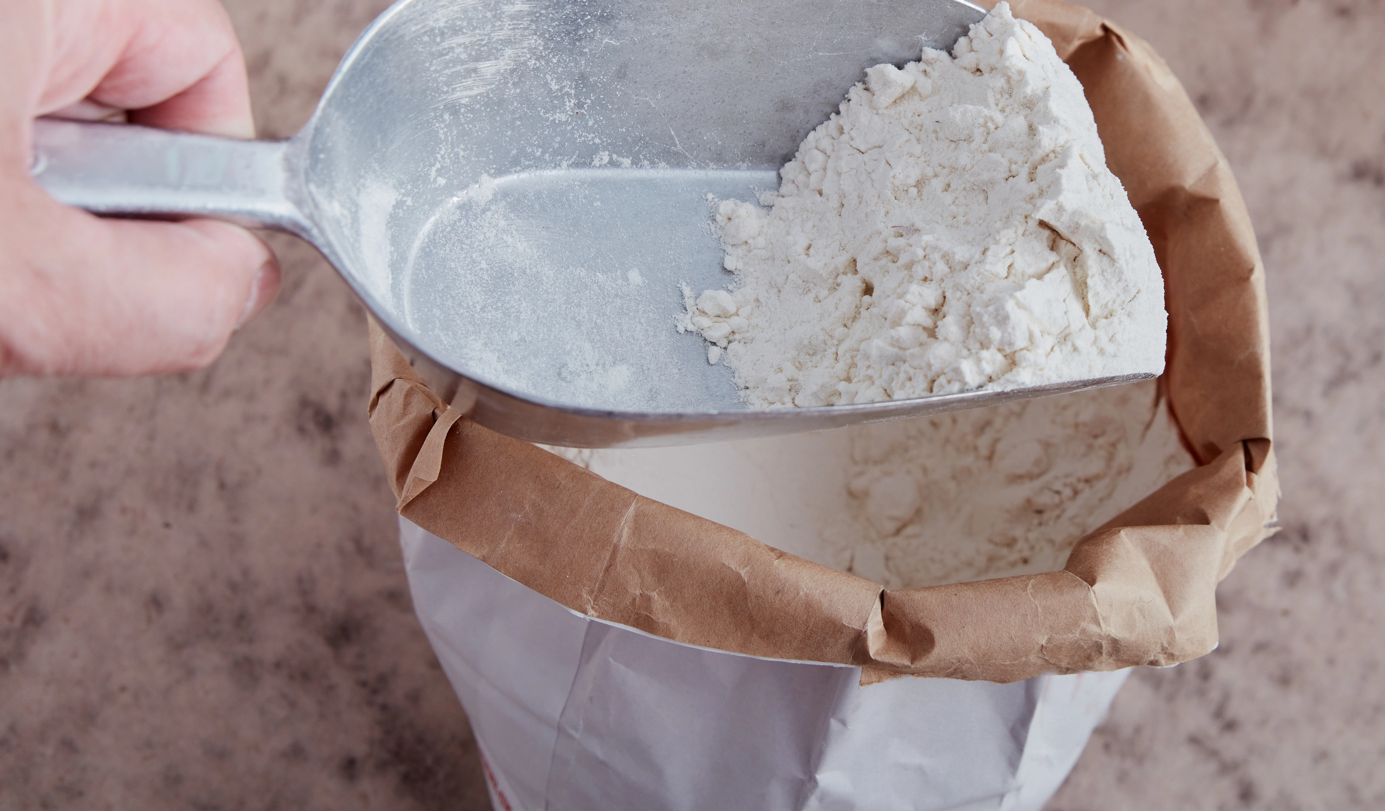 White flour on flour shovel. Opened bag on brownish surface. High point of view.